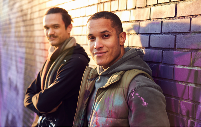 Two men next to each other leaning against brick wall