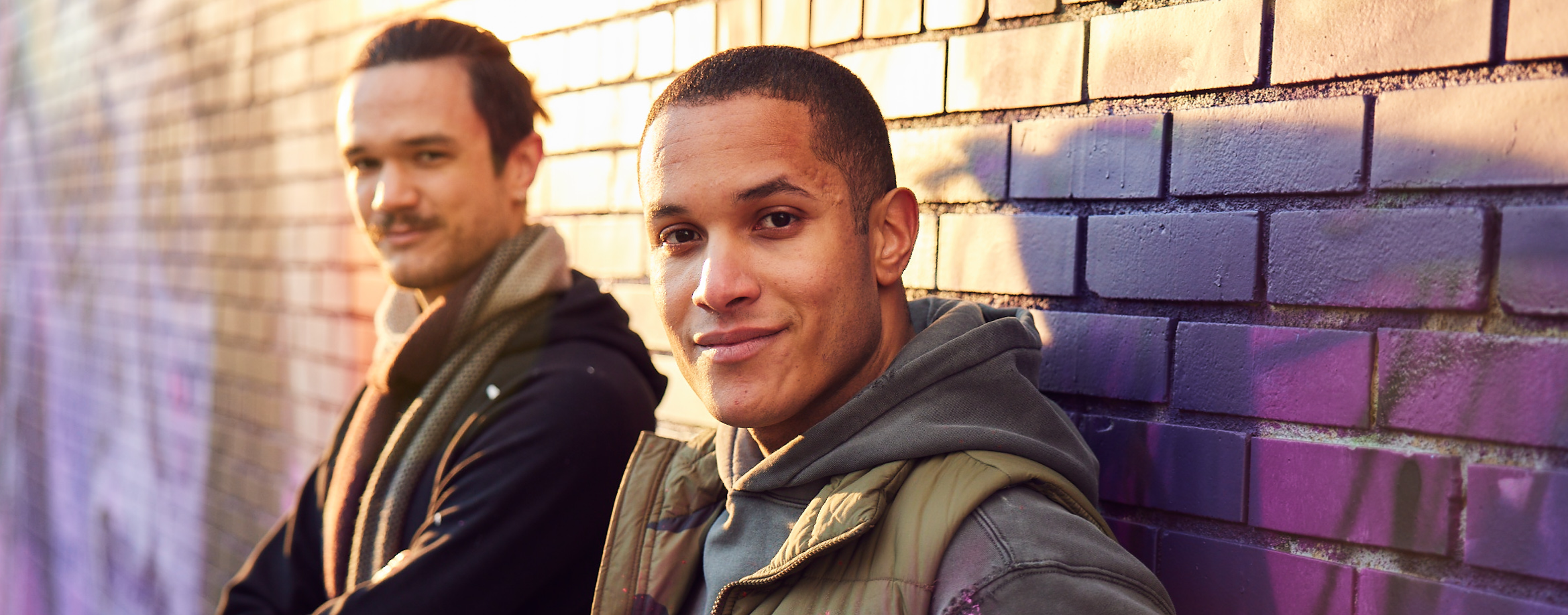 Two men next to each other leaning against brick wall